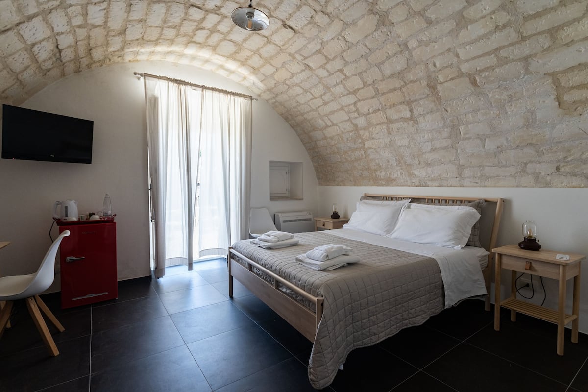 A bedroom with a stone vaulted ceiling, a bed with white linens, a red cabinet, a TV mounted on the wall, and a wooden nightstand with a lamp.