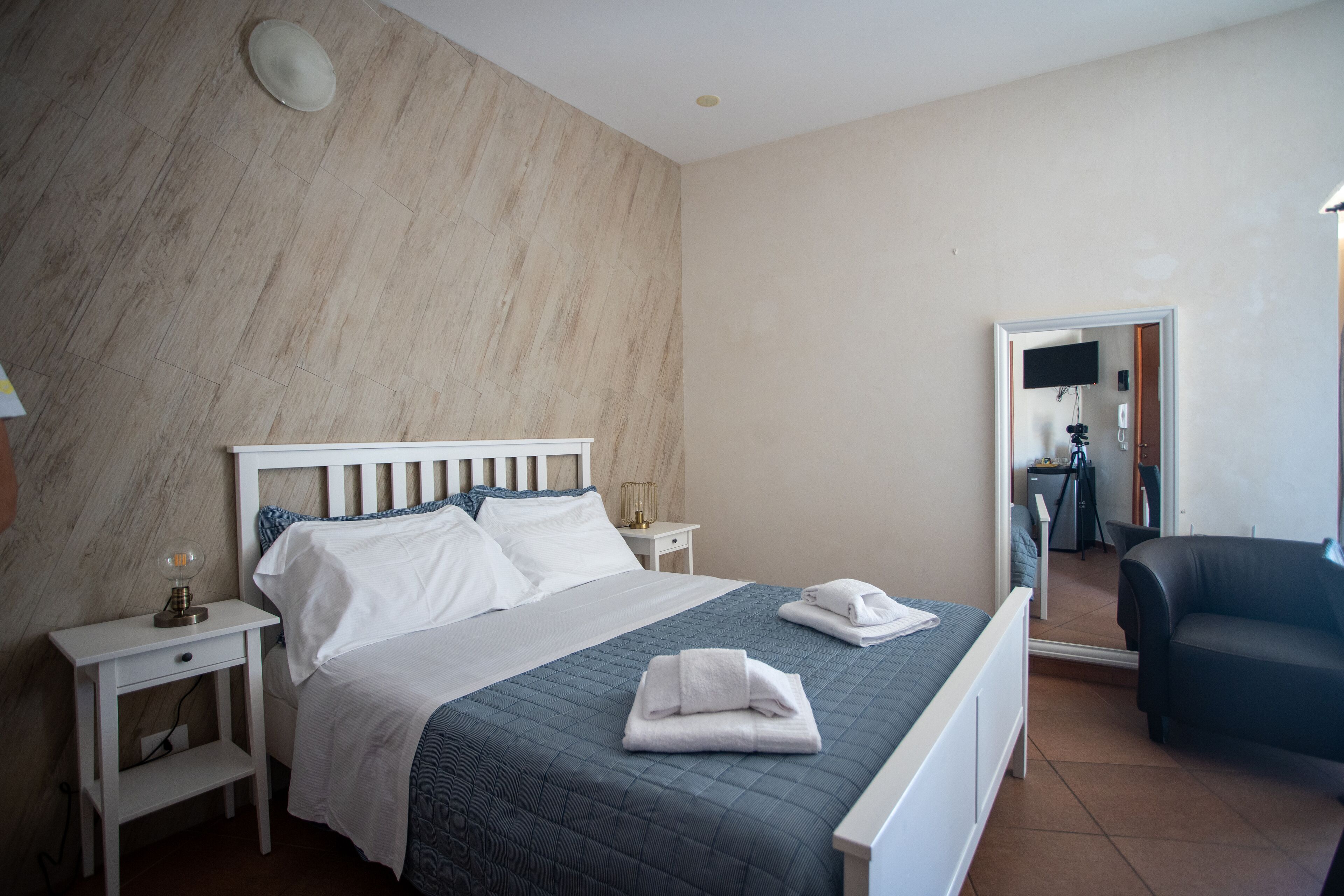 Una cama bien hecha con sábanas blancas y colcha azul, una mesita de noche con lámpara, y una silla en una habitación con suelo de baldosas y un espejo grande.