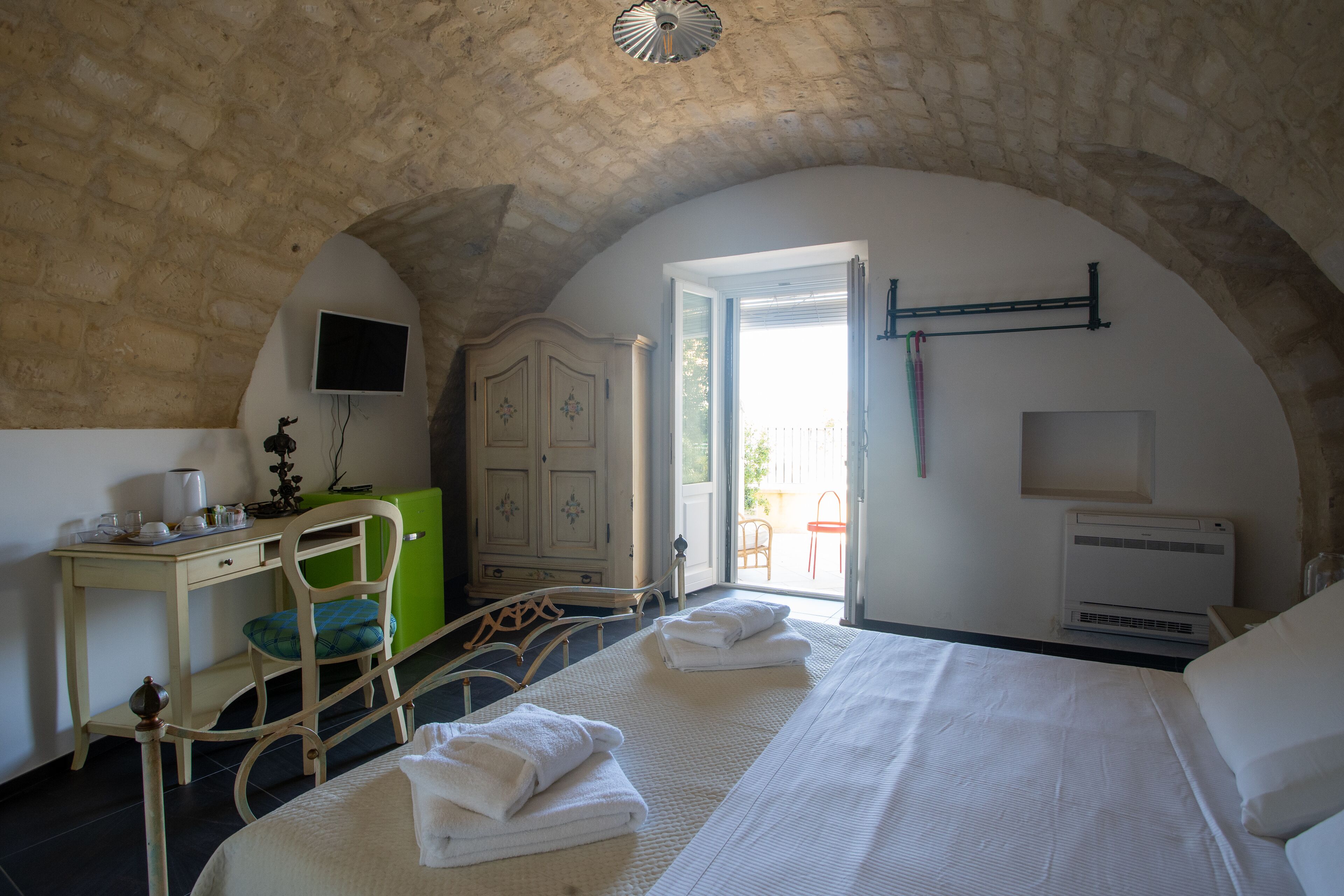 A bedroom with a stone ceiling, a bed with white linens, a desk with a computer, a green chair, and a wardrobe.
