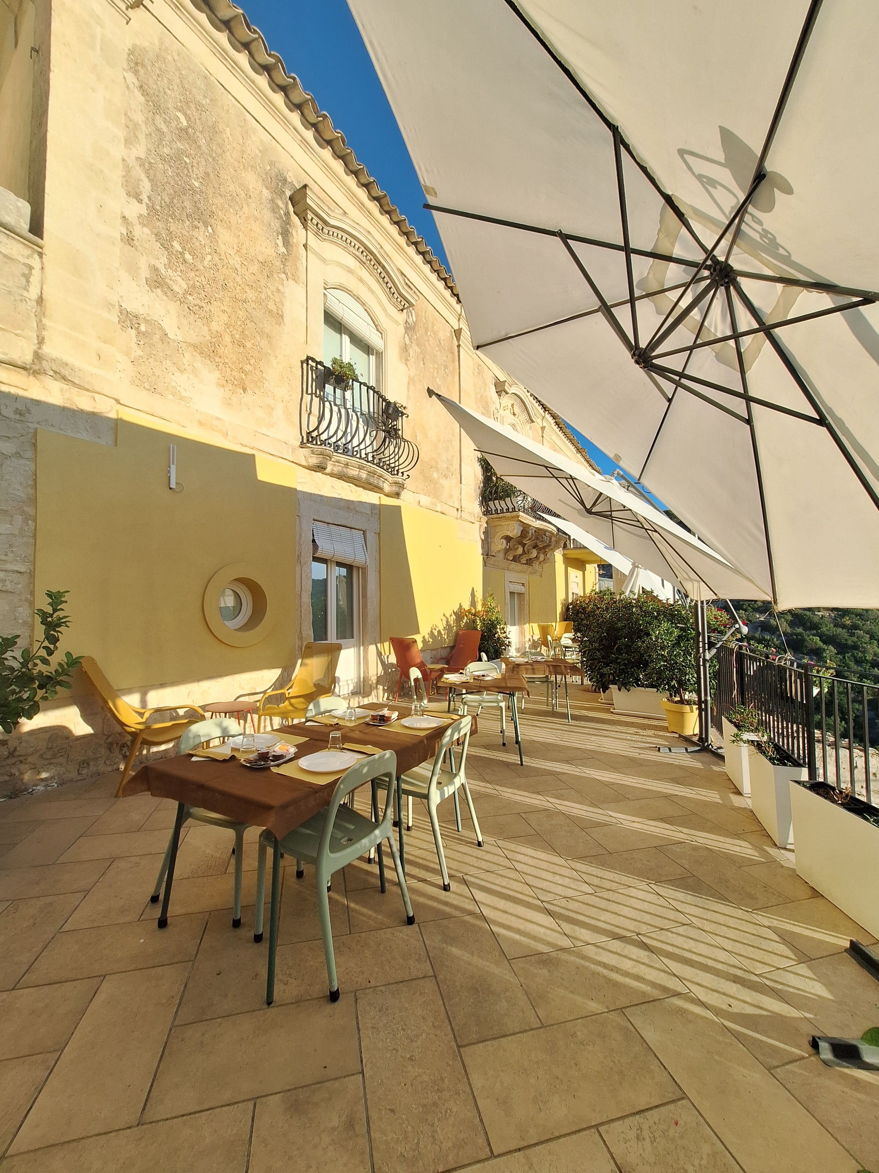 An outdoor seating area with tables and chairs, a building with a balcony, and a large umbrella providing shade.