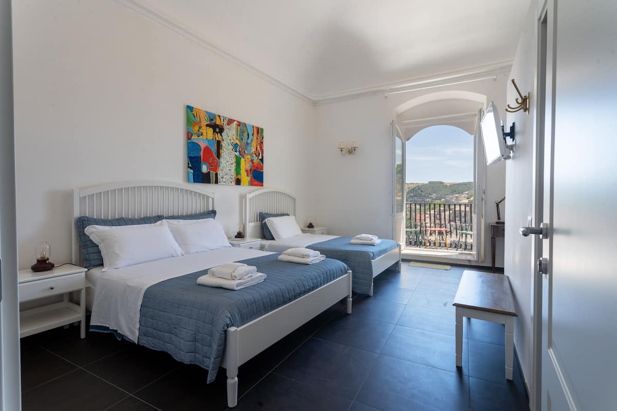 A bedroom with two beds, a colorful abstract painting, a balcony with a view, and a small table.
