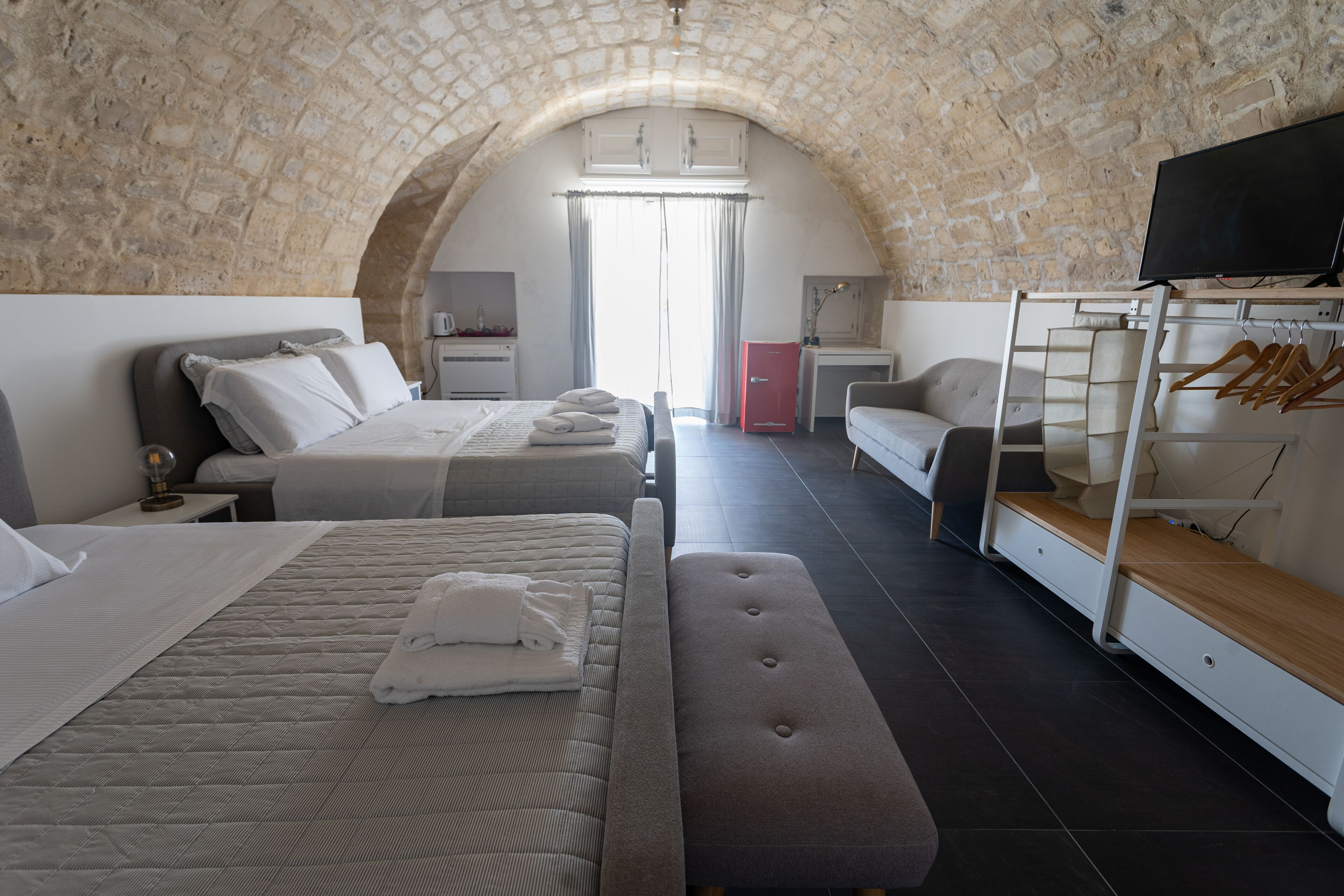 Habitación de hotel con techo de piedra abovedado, una cama con almohada y toalla doblada, un banco, un televisor, un armario y un perchero.