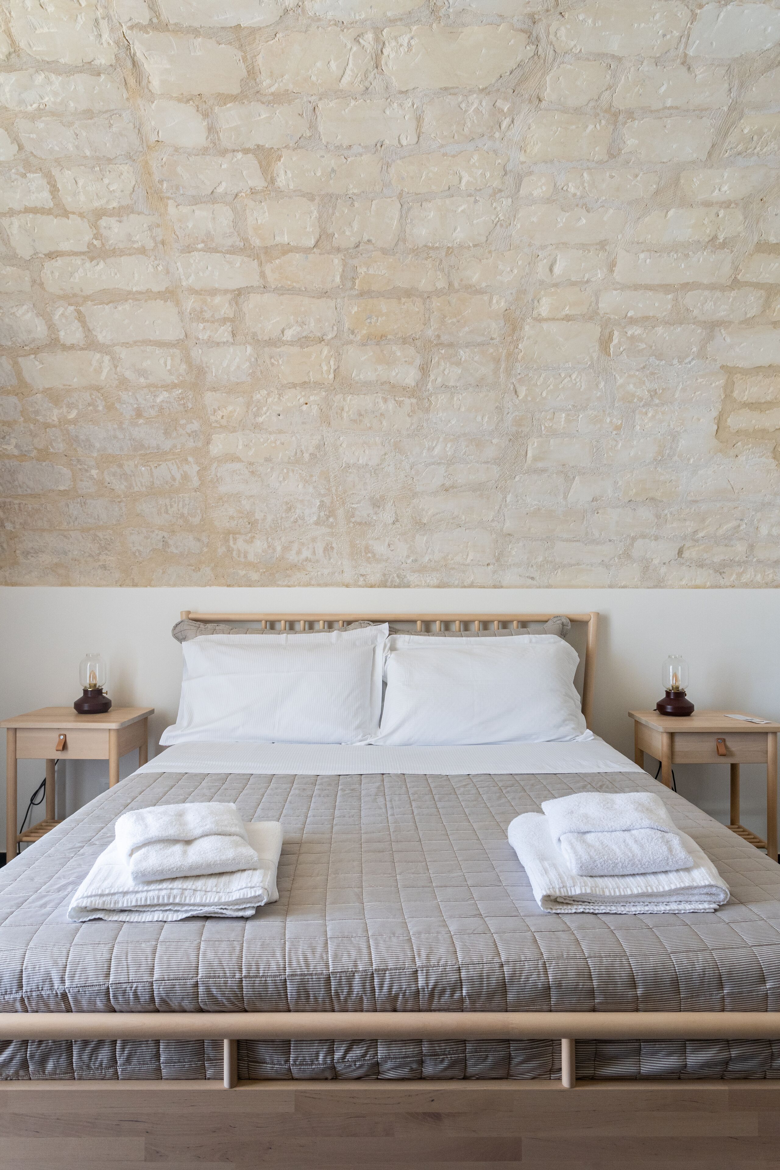 Un letto con struttura in legno, due cuscini bianchi e una coperta trapuntata grigia. Ci sono due comodini con lampade e un piccolo cassetto. La parete dietro il letto è rivestita di mattoni color beige.