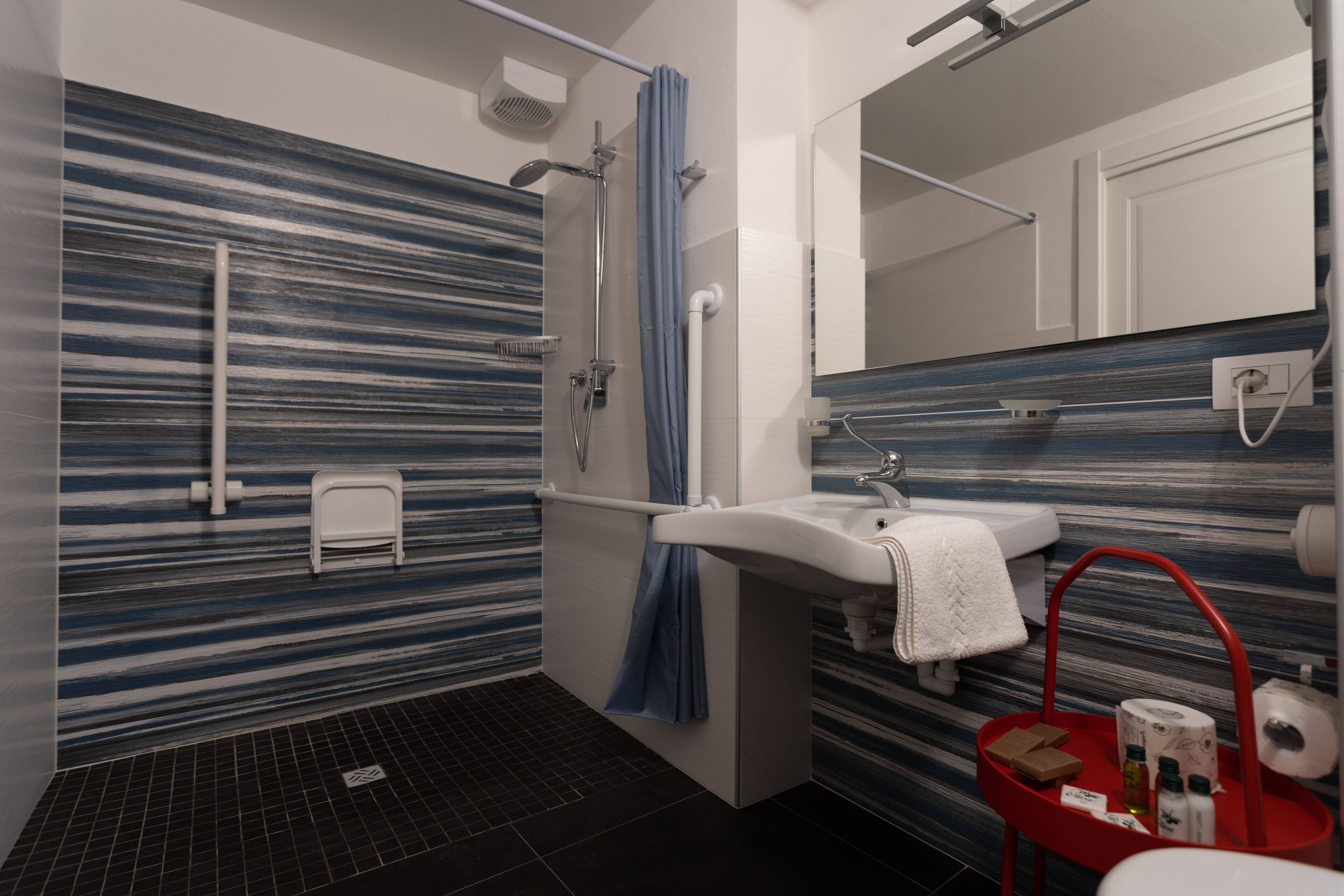 A modern bathroom with a striped tile wall, a shower, a sink, and a red tray with toiletries.