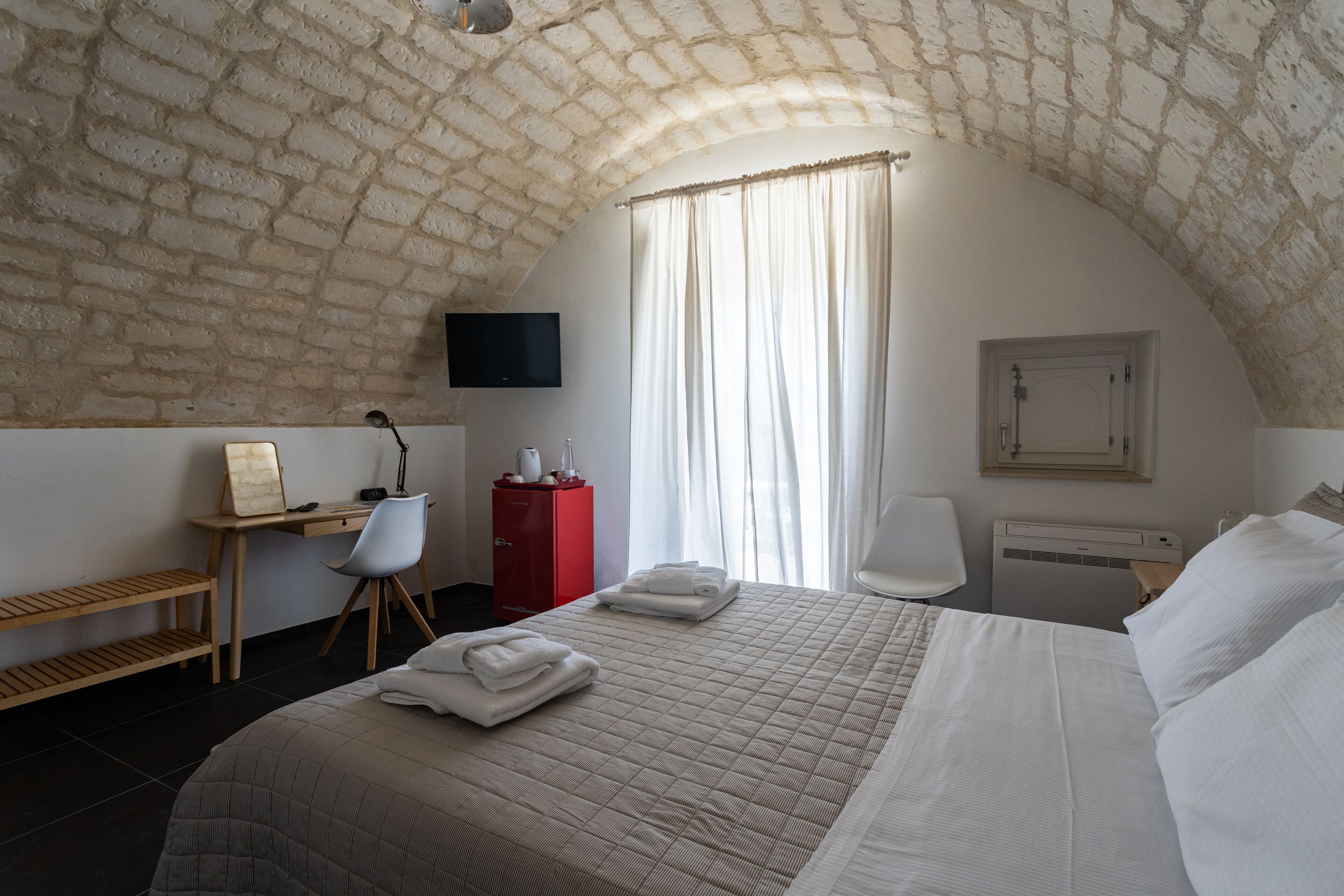 A bedroom with a brick ceiling, a bed with neatly folded towels, a desk with a chair, a red cabinet, a TV, and a window with curtains.