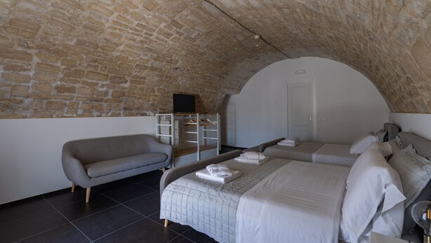 Un dormitorio con techo de piedra, una cama con ropa de cama blanca, un sofá gris y un televisor pequeño sobre una estantería.