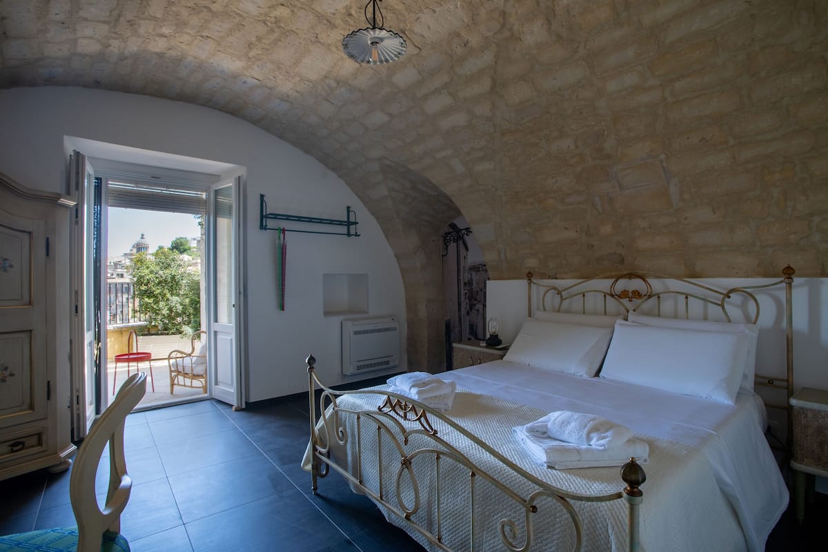 A bedroom with a bed, a chair, a radiator, and an open door leading to a balcony.