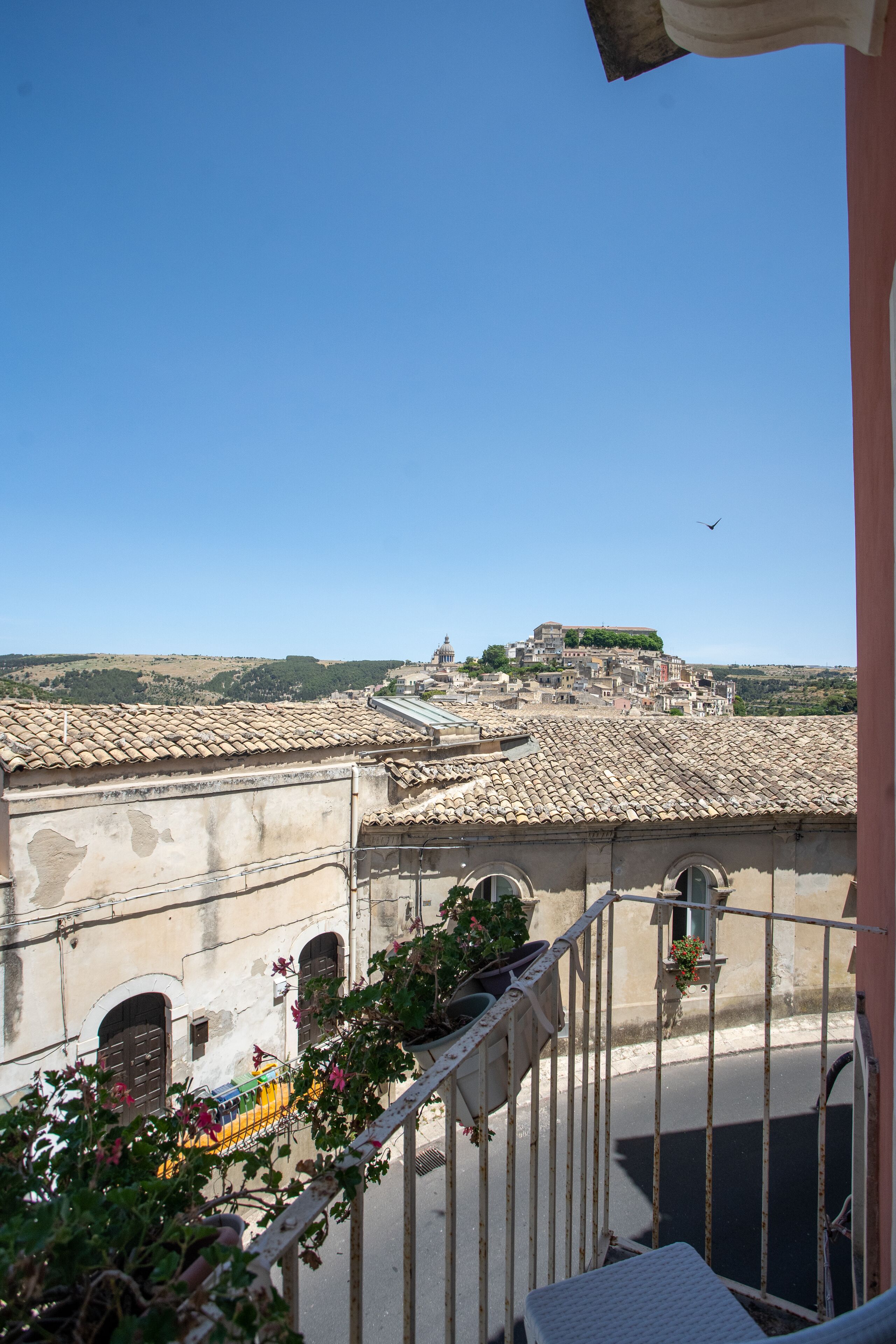 A balcony with a view of terracotta-roofed buildings and a distant hill.