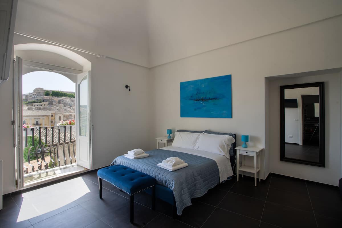 A bedroom with a bed, a bench, a nightstand, a painting, and a balcony with a view of buildings.