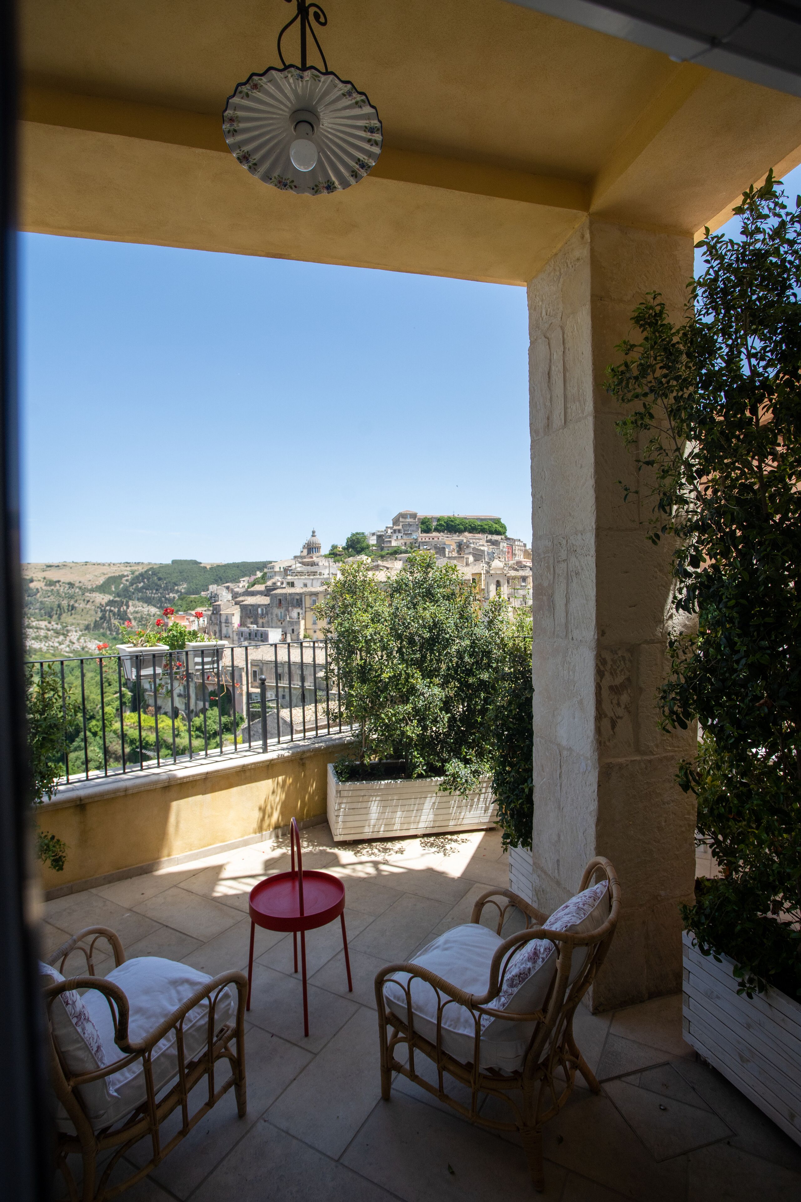 Un balcon agrémenté de fauteuils en osier et d’une petite table, d’où l’on peut admirer un panorama urbain avec ses bâtiments et sa verdure.