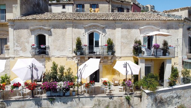 Un edificio con varios balcones, mobiliario exterior con sombrillas y arreglos florales.