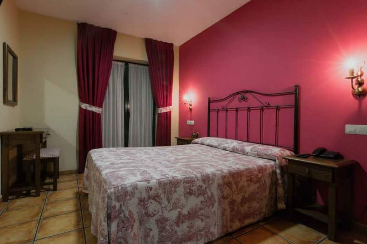 Un dormitorio con cama, mesita de noche, escritorio, silla y ventana con cortinas.