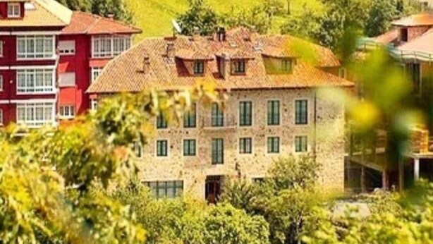 A multi-story building with a stone facade and multiple windows, surrounded by greenery and other buildings. - Hotel Villa de Cabrales (Cabrales)