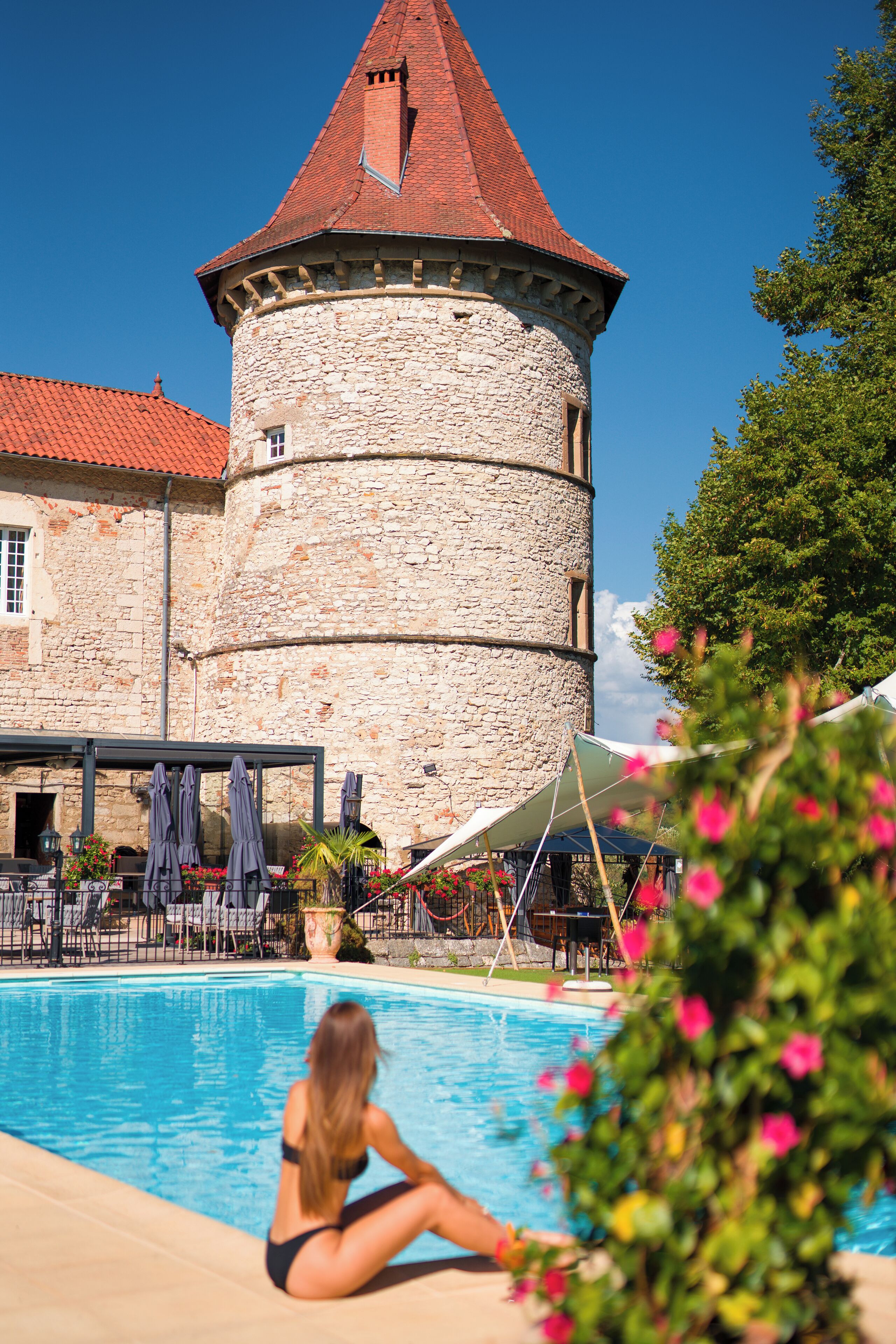 Une tour en pierre surmontée d’un toit rouge, une piscine et un espace extérieur aménagé avec des parasols.