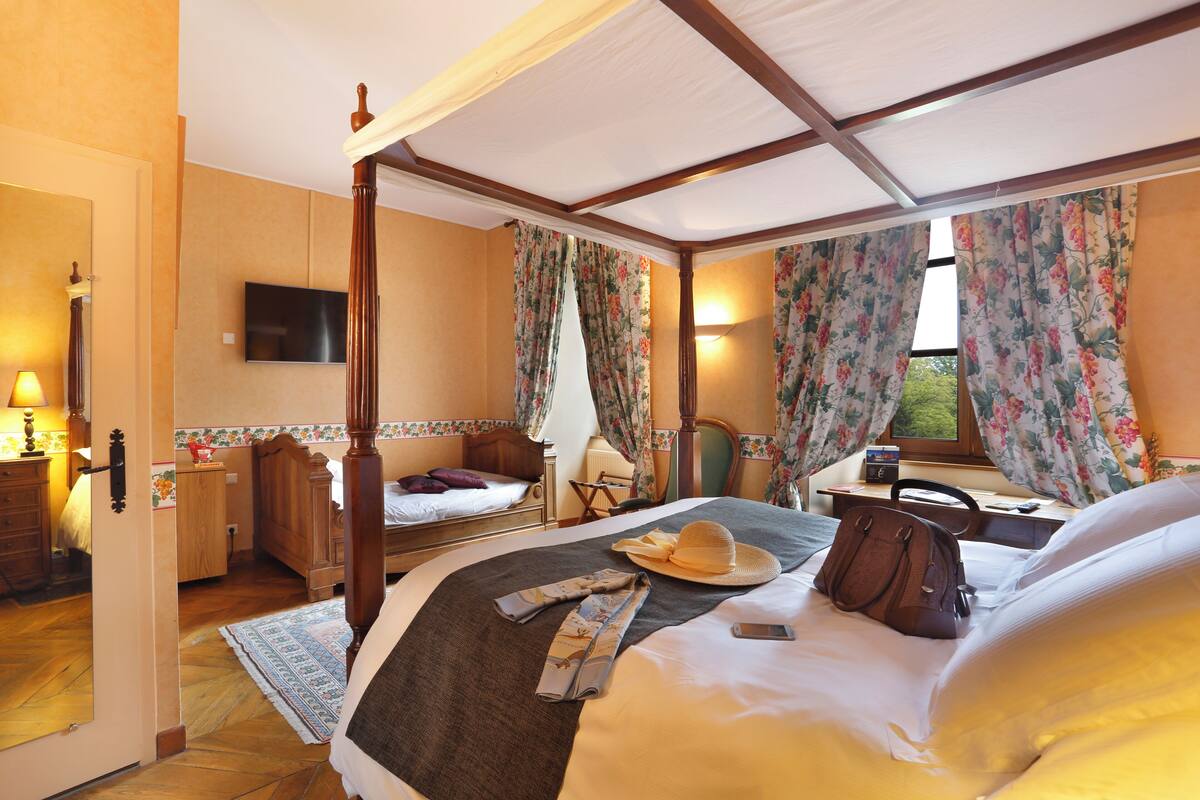 Un lit à baldaquin, une table de chevet avec une lampe, une chaise, un bureau avec un ordinateur et un téléviseur fixé au mur.