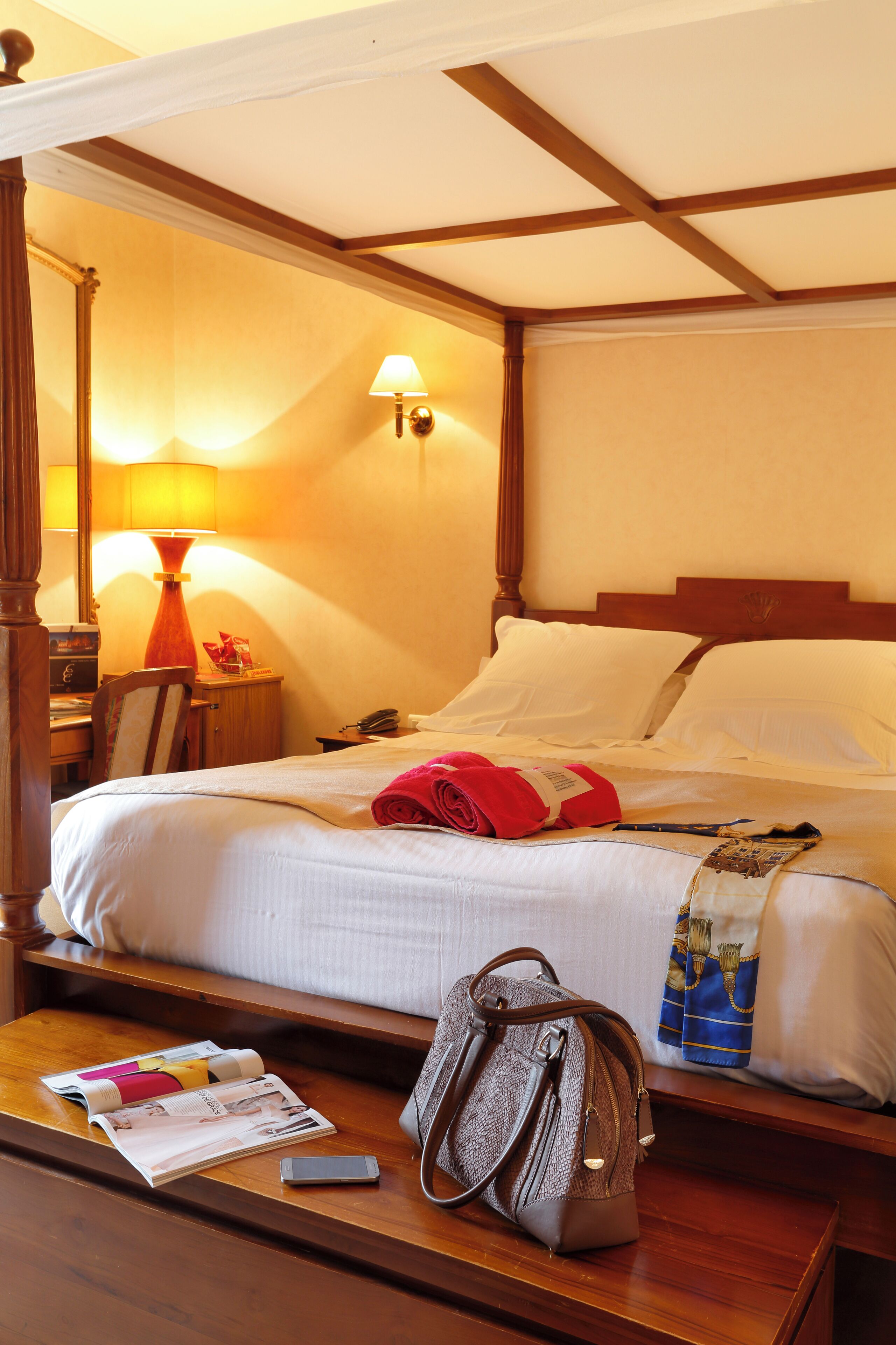 A hotel room with a bed, a nightstand, a lamp, a chair, a table with a magazine and a handbag, and a wall-mounted lamp.