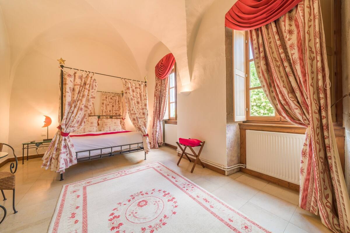 Une chambre à coucher avec un lit à baldaquin, un fauteuil rouge, un plancher en bois et une grande fenêtre avec des rideaux.
