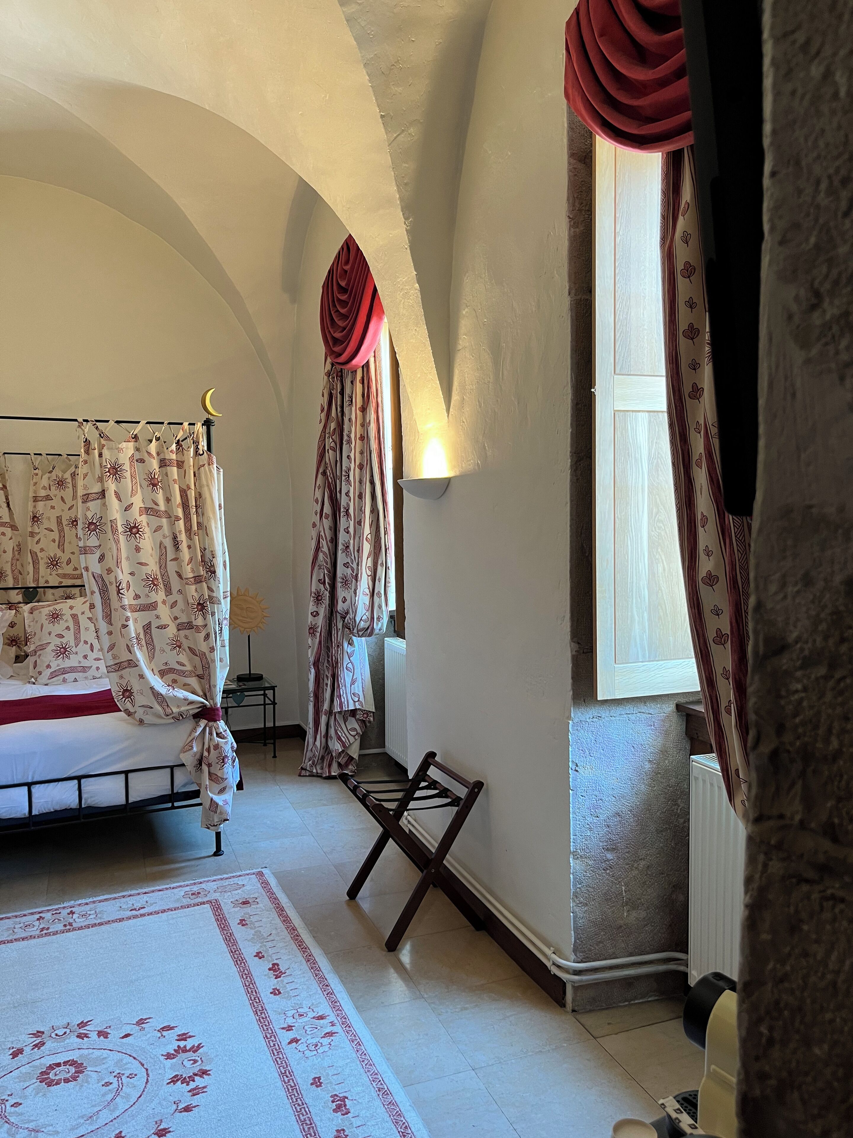 A bedroom with a bed, a nightstand, a chair, a window with curtains, and a wall-mounted light.