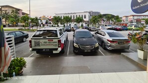Un parking avec plusieurs voitures, un fourgon blanc sur lequel figure un numéro de téléphone, et un bâtiment avec une enseigne indiquant « Surau Al-Hikmah Kuta Wisma ».