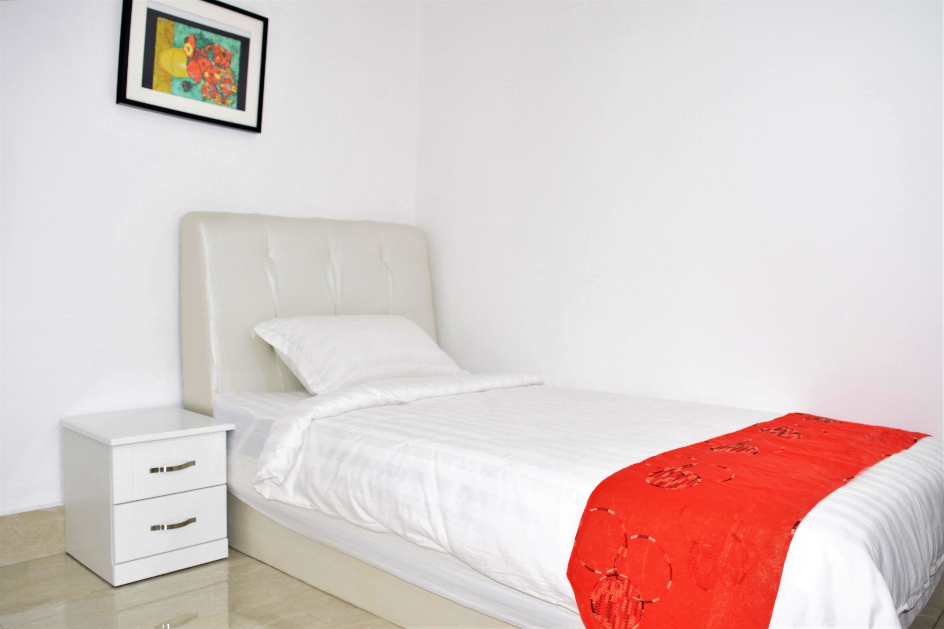 Un letto rifatto con cura, con testiera bianca e coperta rossa, un comodino con cassetti e un quadro appeso al muro.