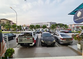 A parking lot with several cars, a white van with a contact number, and a building with a sign that reads "Surau Al-Hikmah Kuta Wisma".