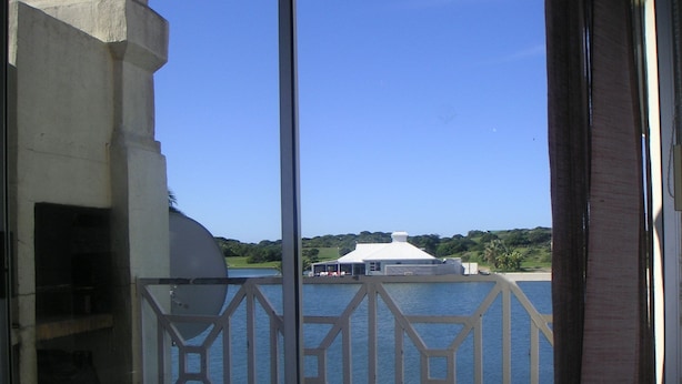 A view through a window with a geometric metal railing, overlooking a body of water and a white building with a sloped roof.