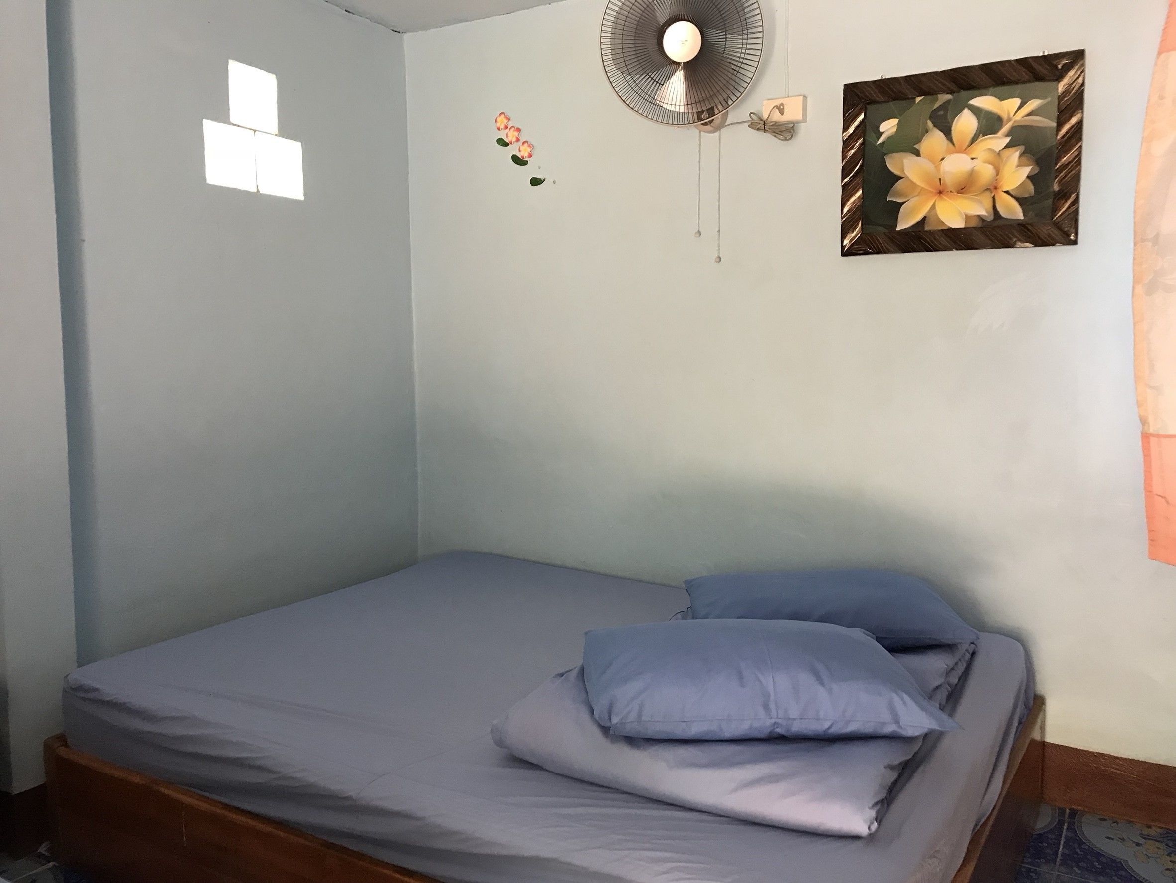 Een enkel bed met een grijze matras en twee kussens, een ventilator bevestigd aan de muur, een lijst met bloemen erin, en een klein raam met een witte rand.