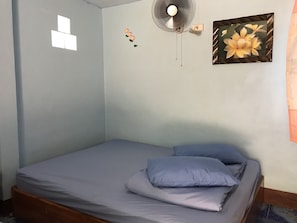 Uma cama de solteiro com colchão cinzento e duas almofadas, um ventilador fixado na parede, um quadro com flores e uma pequena janela com moldura branca.