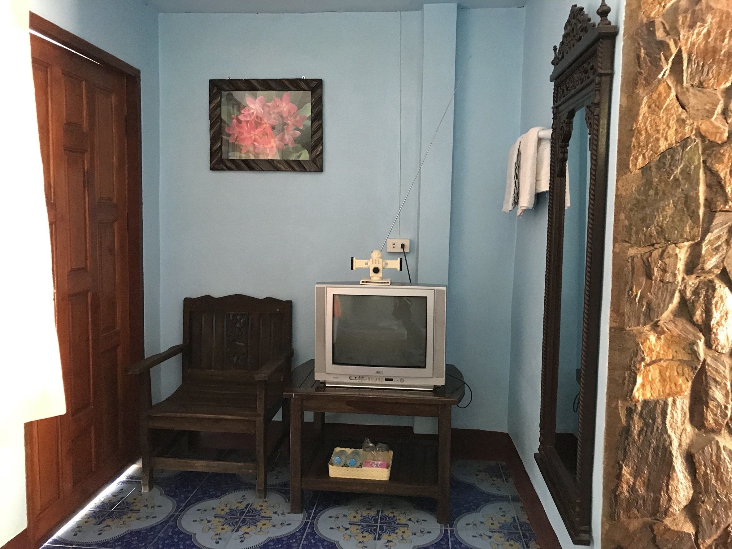 A room with a wooden door, a framed picture on the wall, a television on a stand, a stone wall, and a tiled floor.