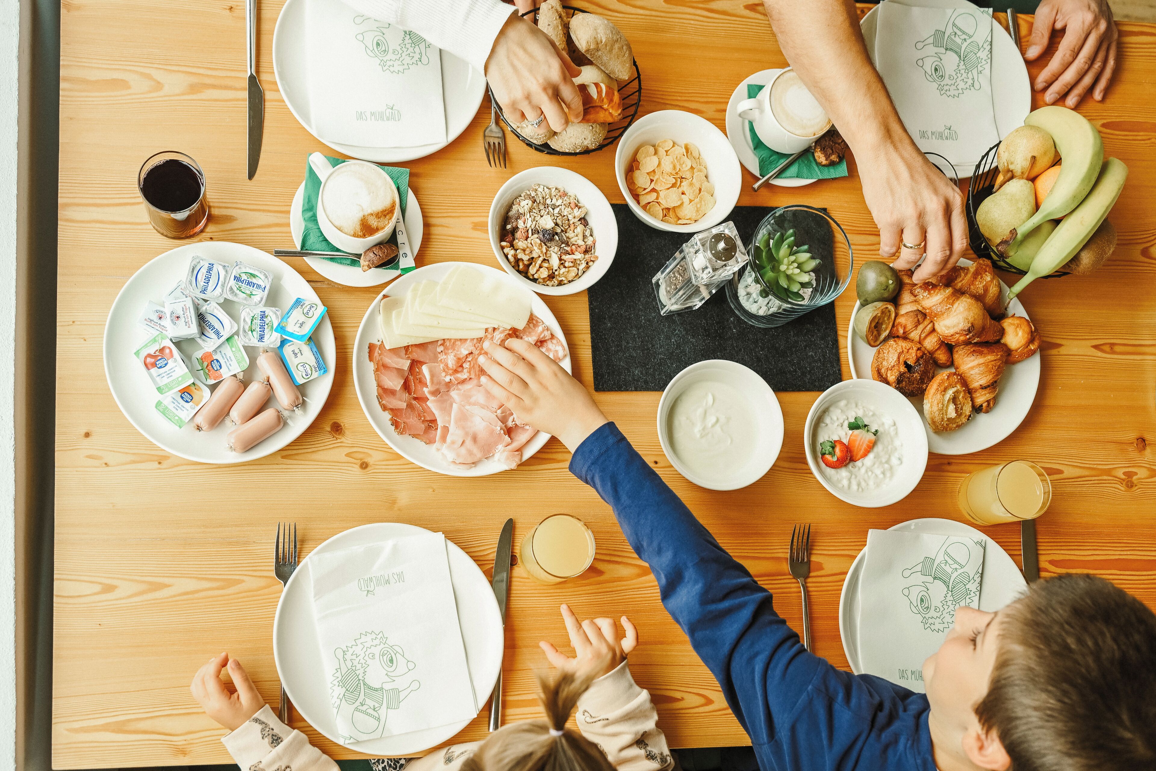A wooden table with various food items and plates, utensils, and a napkin with a drawing.