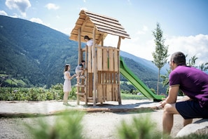 A playground with a wooden structure and a slide, set against a mountainous backdrop. - Das Mühlwald Quality Time Family Resort (Naz Sciaves)