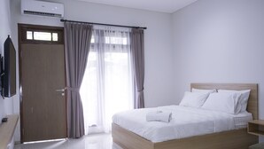 A modern hotel room with a wooden bed, white bedding, a flat-screen TV mounted on the wall, and a window with curtains. - MK House Tendean (Jakarta)