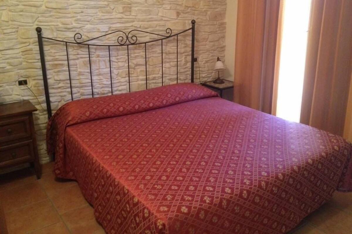 Ein Schlafzimmer mit Steinwand, Metallbettgestell, rot gemustertem Bettbezug, Nachttisch mit Lampe und einem Fenster mit Vorhängen.