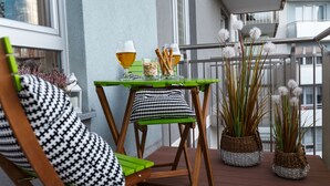 Ein Balkon mit einem Holzstuhl, einem kleinen Tisch mit einem Bierglas und zwei Topfpflanzen.