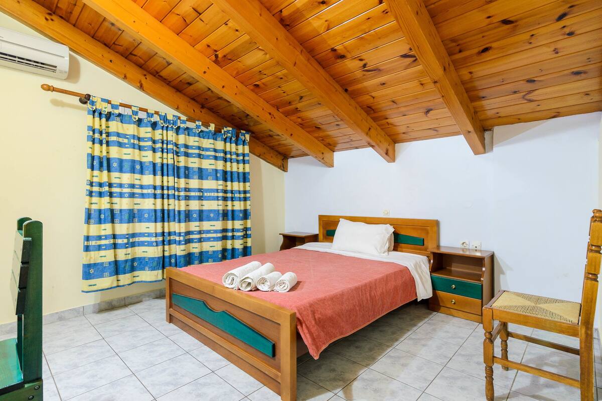 A bedroom with a wooden ceiling, a bed with a red blanket, a wooden nightstand, a chair, and a curtain with a blue and yellow pattern.