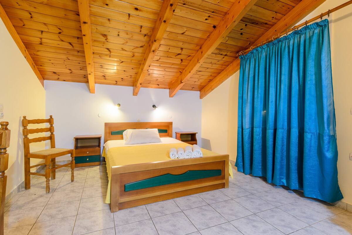 A bedroom with a wooden ceiling, a bed with a green headboard, a wooden chair, a nightstand, and a blue curtain.