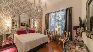 Une chambre d’hôtel avec un grand lit, un lustre, un miroir, un bureau avec une lampe et une vue sur l’extérieur par une fenêtre avec des rideaux.
