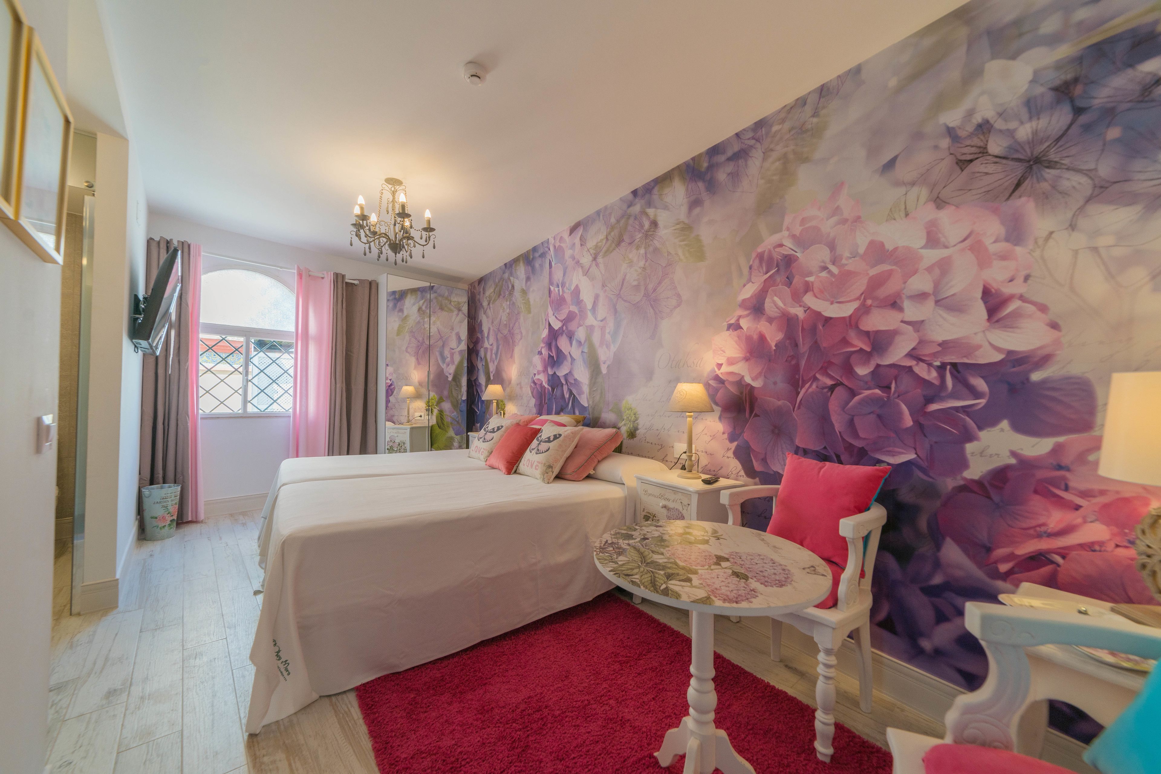 Un dormitorio con dos camas, alfombra roja, mural floral, candelabro y una pequeña mesa con una silla.