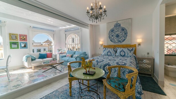A spacious room with a large bed, a dining table, and a seating area with chairs. There is a chandelier, a wall-mounted lamp, and a decorative piece above the bed. - Rey Don Pedro Luxury Home (Seville)