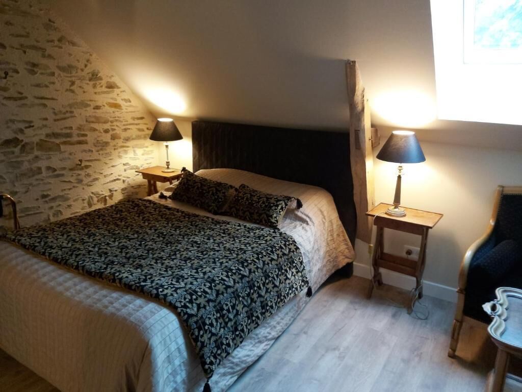 Un dormitorio con pared de piedra, cama con colcha estampada, dos mesitas de noche con lámparas y una silla.