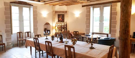 Un comedor con una mesa larga dispuesta para seis personas, rodeada de sillas de madera. Hay dos ventanas que aportan luz natural, un estante en la pared con objetos decorativos y un techo con vigas de madera.