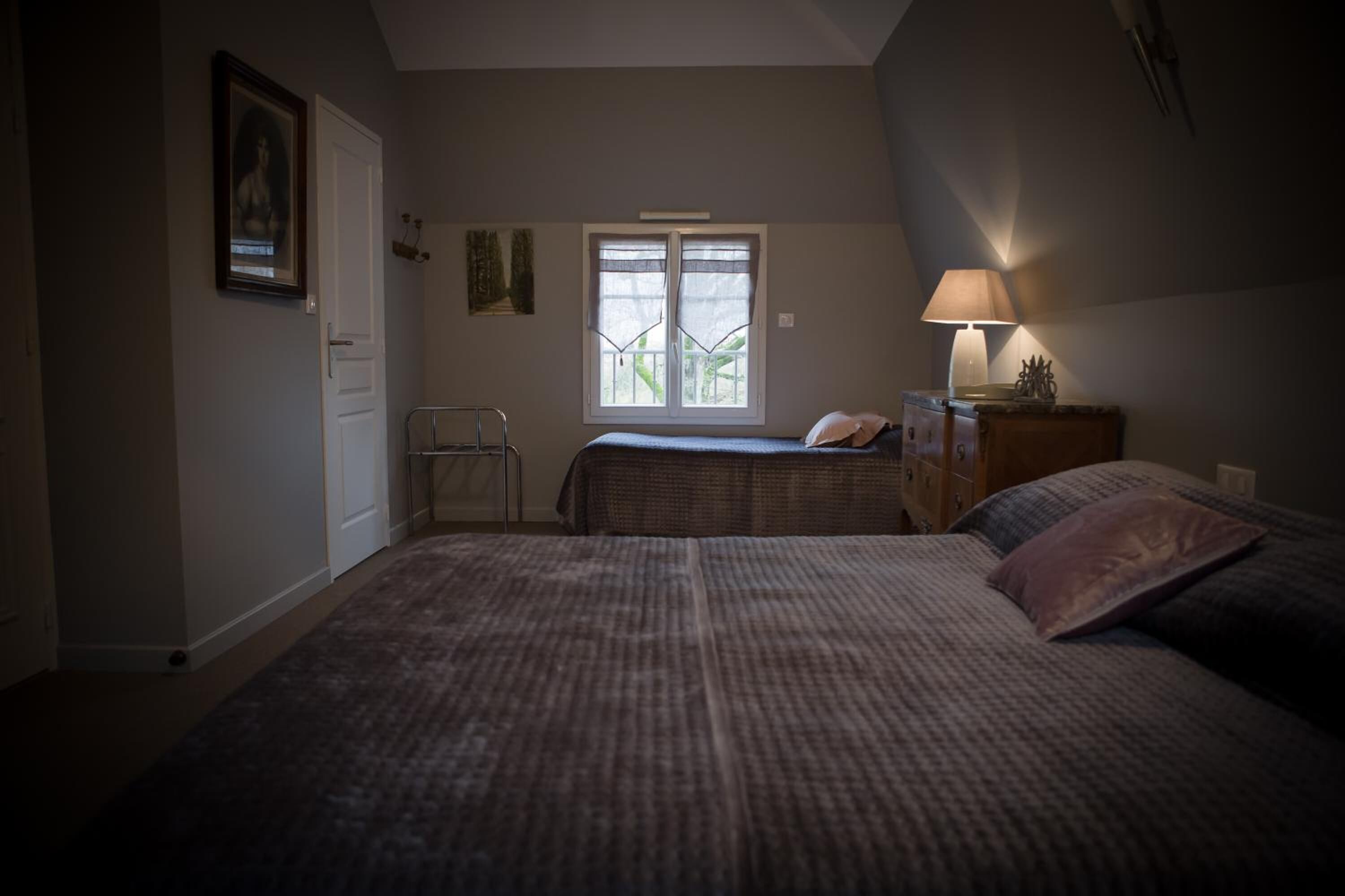 A bedroom with two beds, a window with a view, a bedside table with a lamp, and a door.