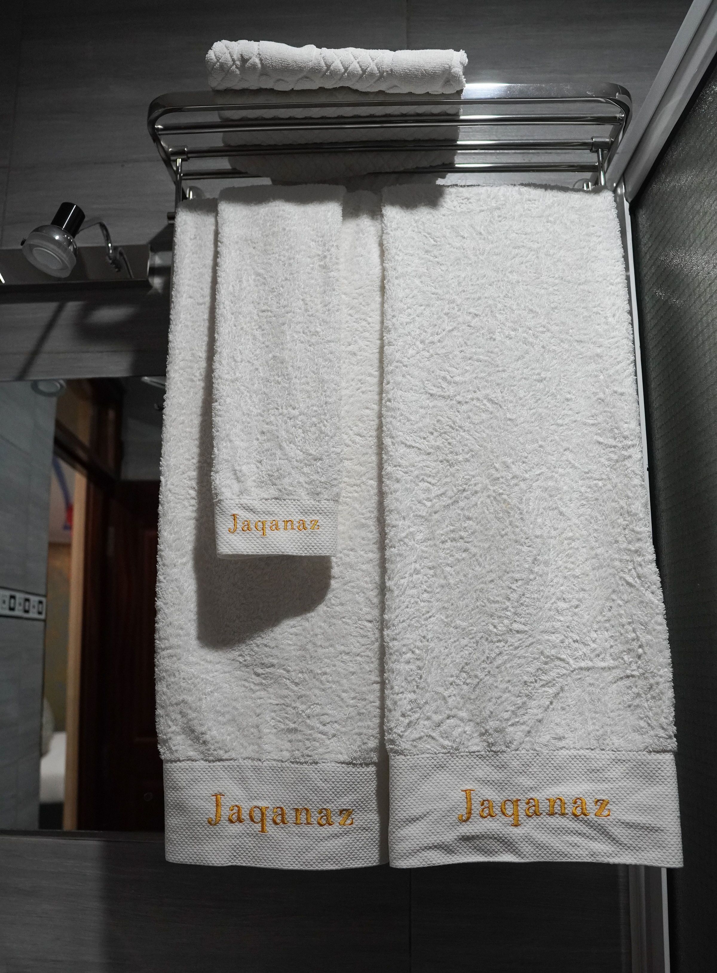 Un baño con dos toallas blancas colgadas en un perchero metálico, cada una con el nombre 'Jaqanaz' bordado en dorado.