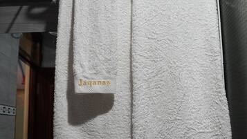 Un baño con dos toallas blancas colgadas en un perchero metálico, cada una con el nombre 'Jaqanaz' bordado en dorado.