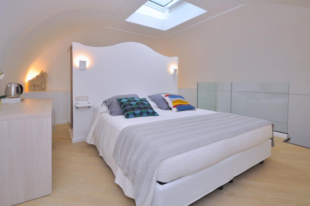 Un dormitorio moderno con tragaluz, una cama con almohadillas estampadas, una mesita de noche y un armario blanco.