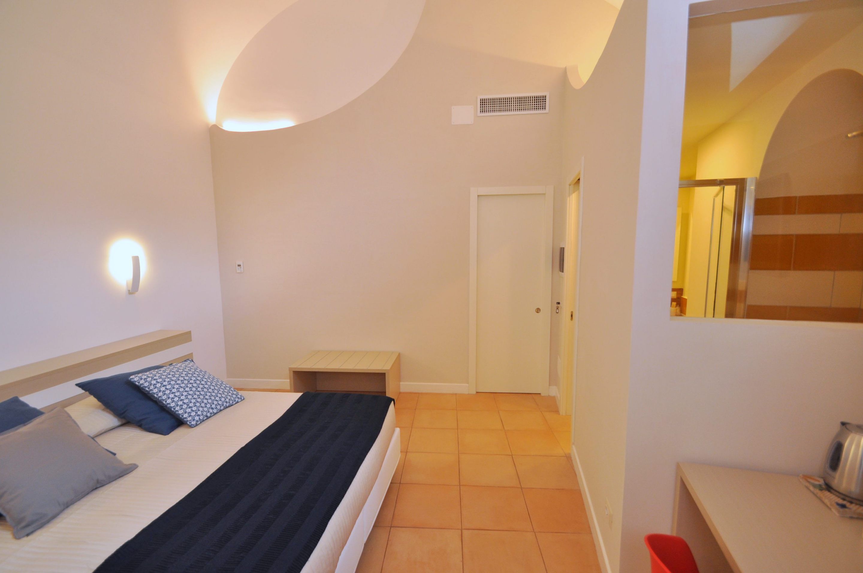 A bedroom with a bed, bedside table, and a small stool. There is a door leading to another room and a mirror on the wall.
