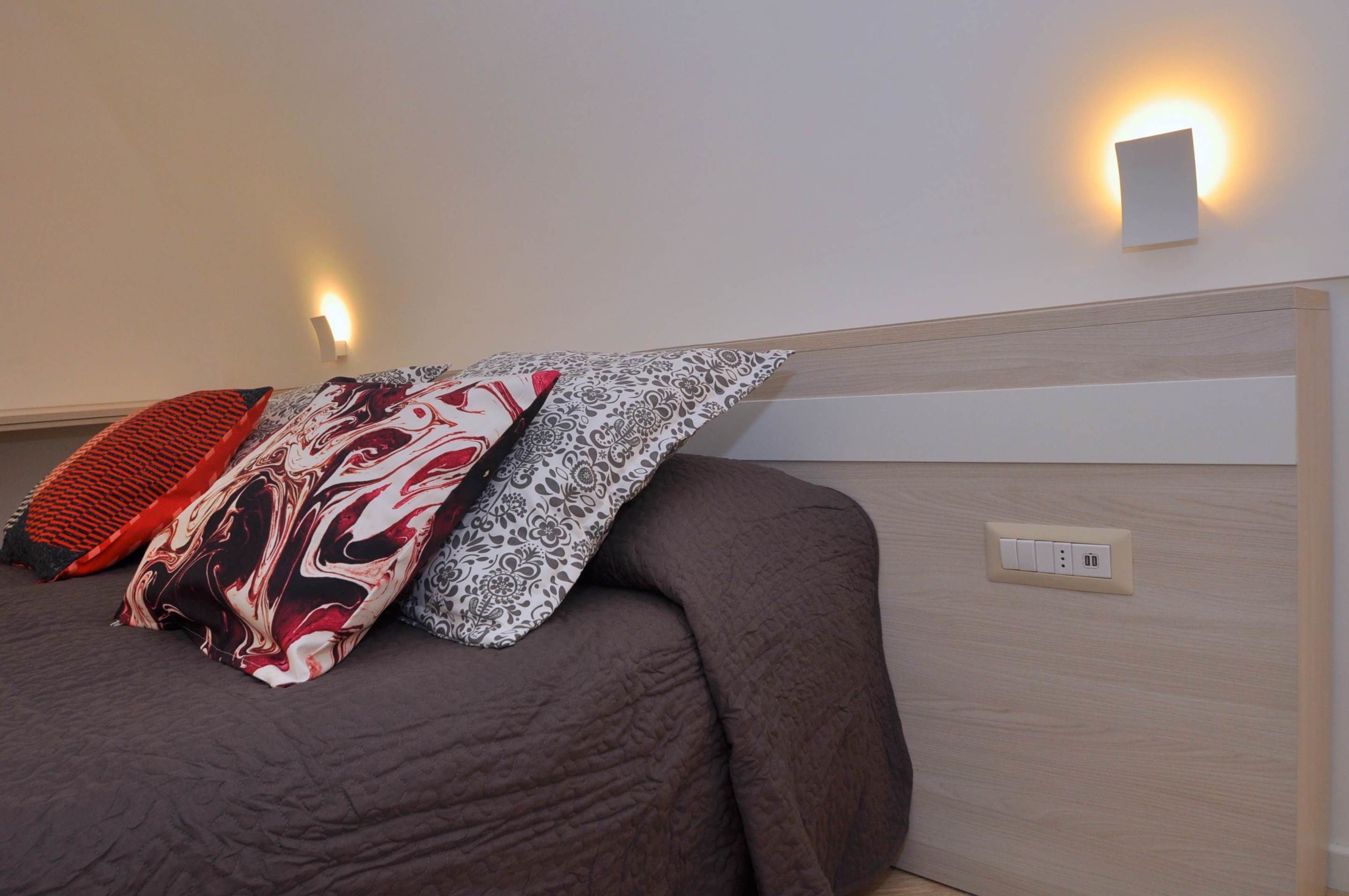 Ein Bett mit einer braunen Bettdecke, gemusterten Kissen, einem hellen Kopfteil und einer Wandleuchte.