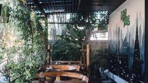 An indoor seating area with wooden furniture, a mural of a city skyline, and lush greenery. - Kristi House (Chiang Mai)