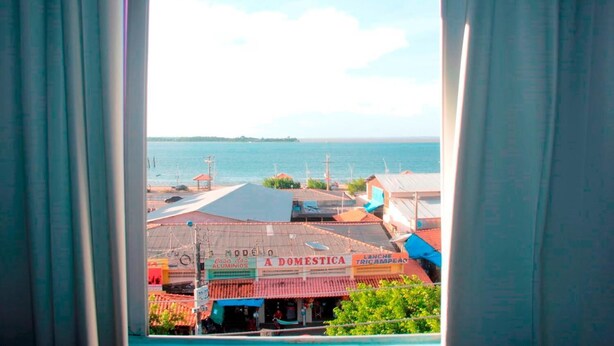 A coastal view from a window with curtains, featuring a building with a sign 'Casa da Aluminio' and 'A Domestica'. - Grand Center Hotel (Santarém)
