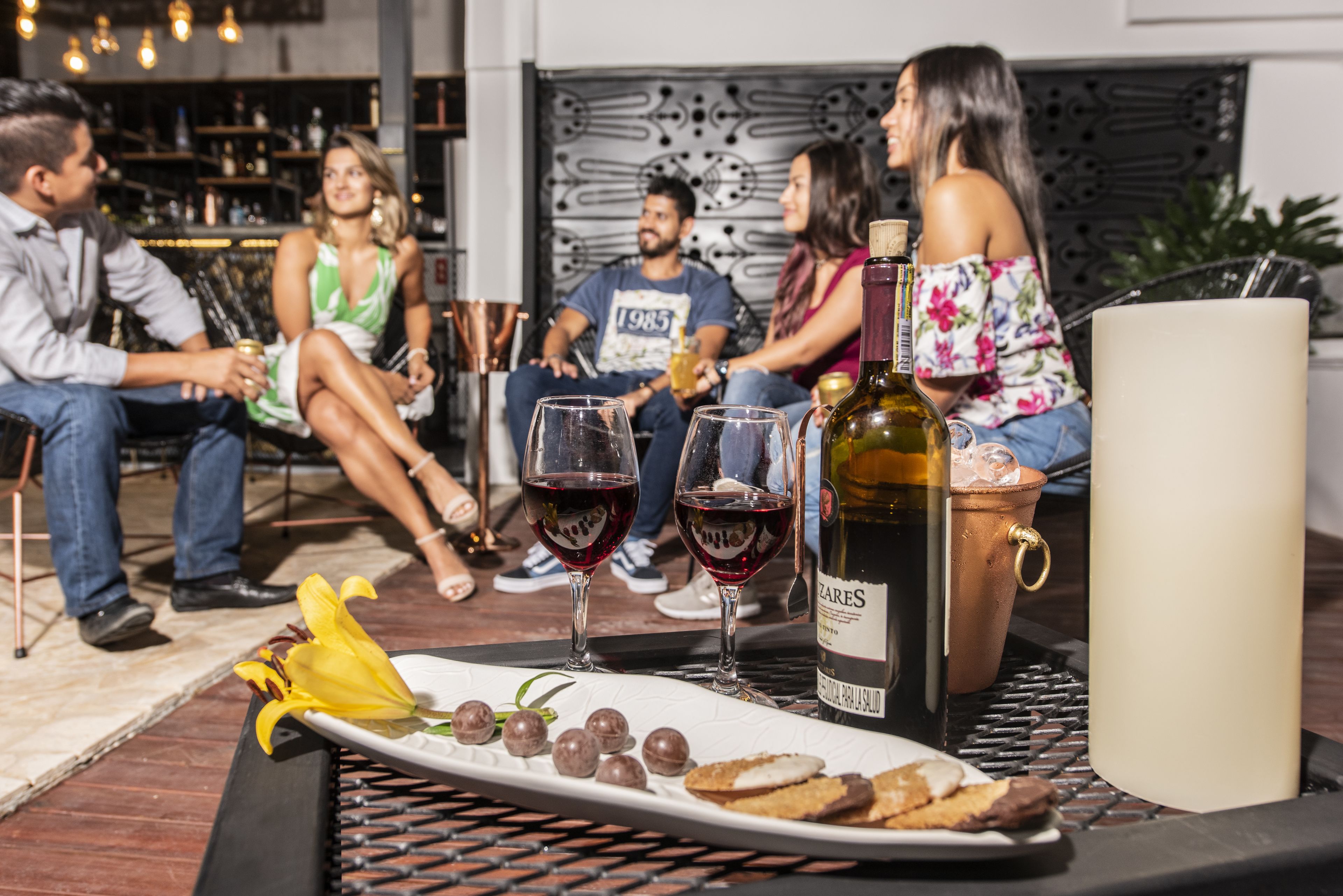 A group of people seated at an outdoor table with wine glasses, a bottle of wine, and a plate of snacks.
